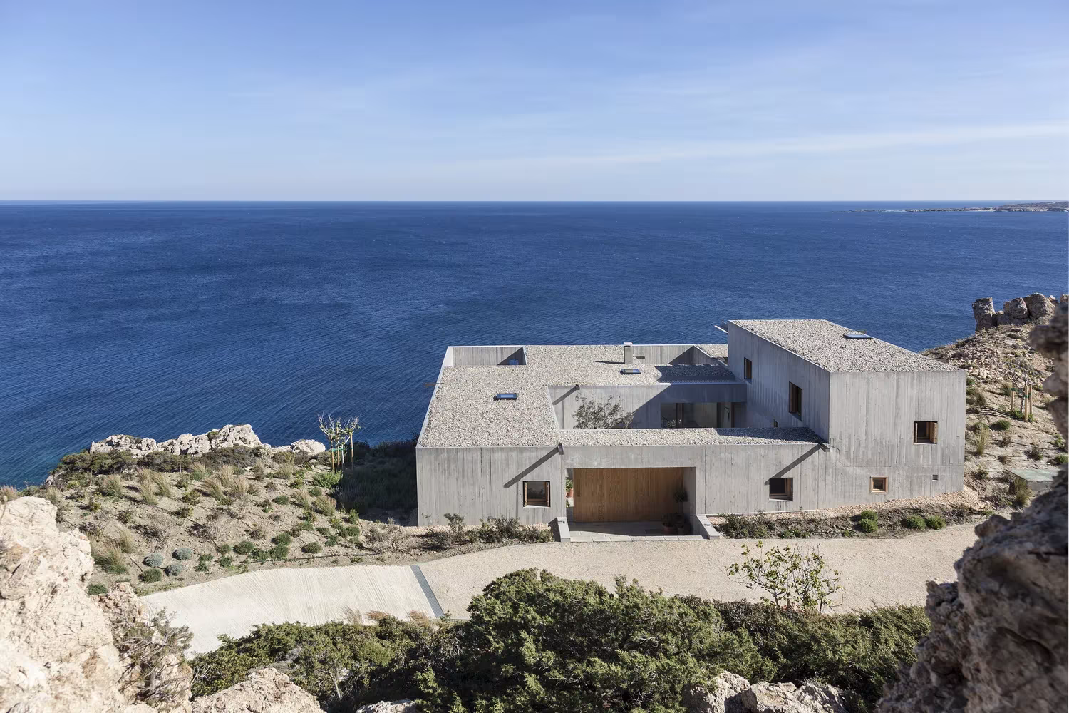 Casa Patio de hormigón visto sobre el acantilado con el mar Egeo al fondo y camino de grava