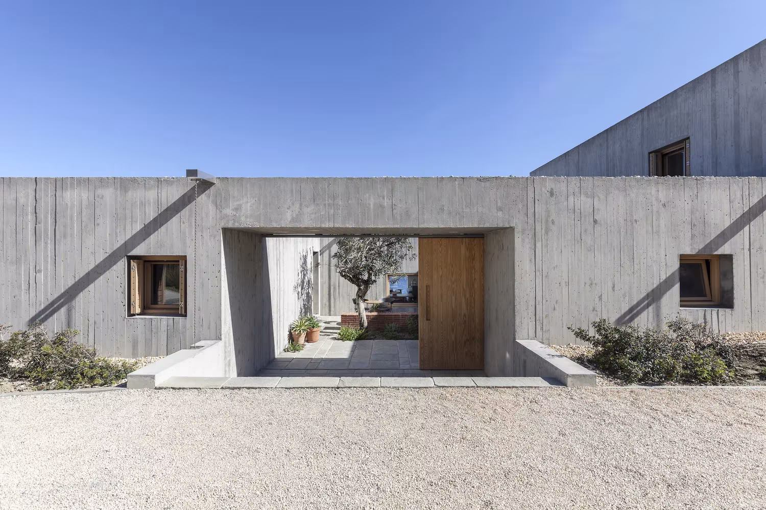Acceso a la Casa Patio por un pórtico hueco en el muro de hormigón encofrado, con puerta de madera