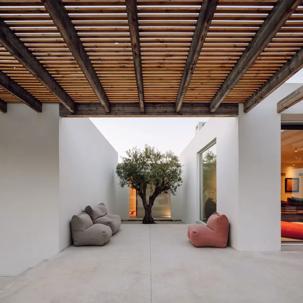 Patio de Comporta 10 bajo pérgola de vigas de madera, con un olivo entre muros blancos y pufs de colores