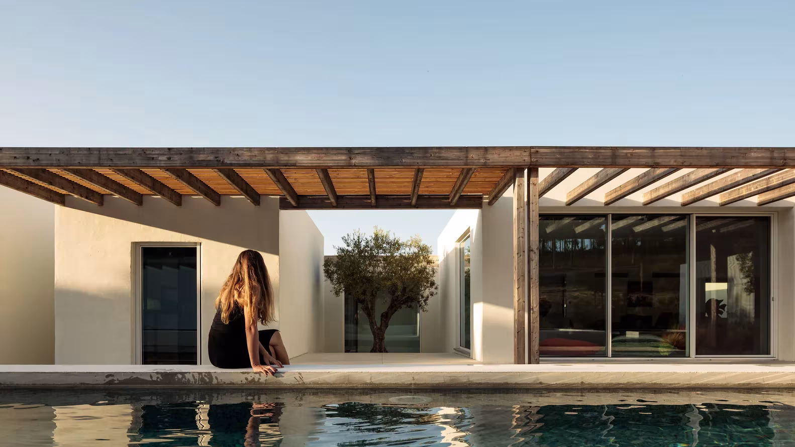 Persona sentada al borde de la piscina frente a la pérgola de madera y los volúmenes blancos de Comporta 10