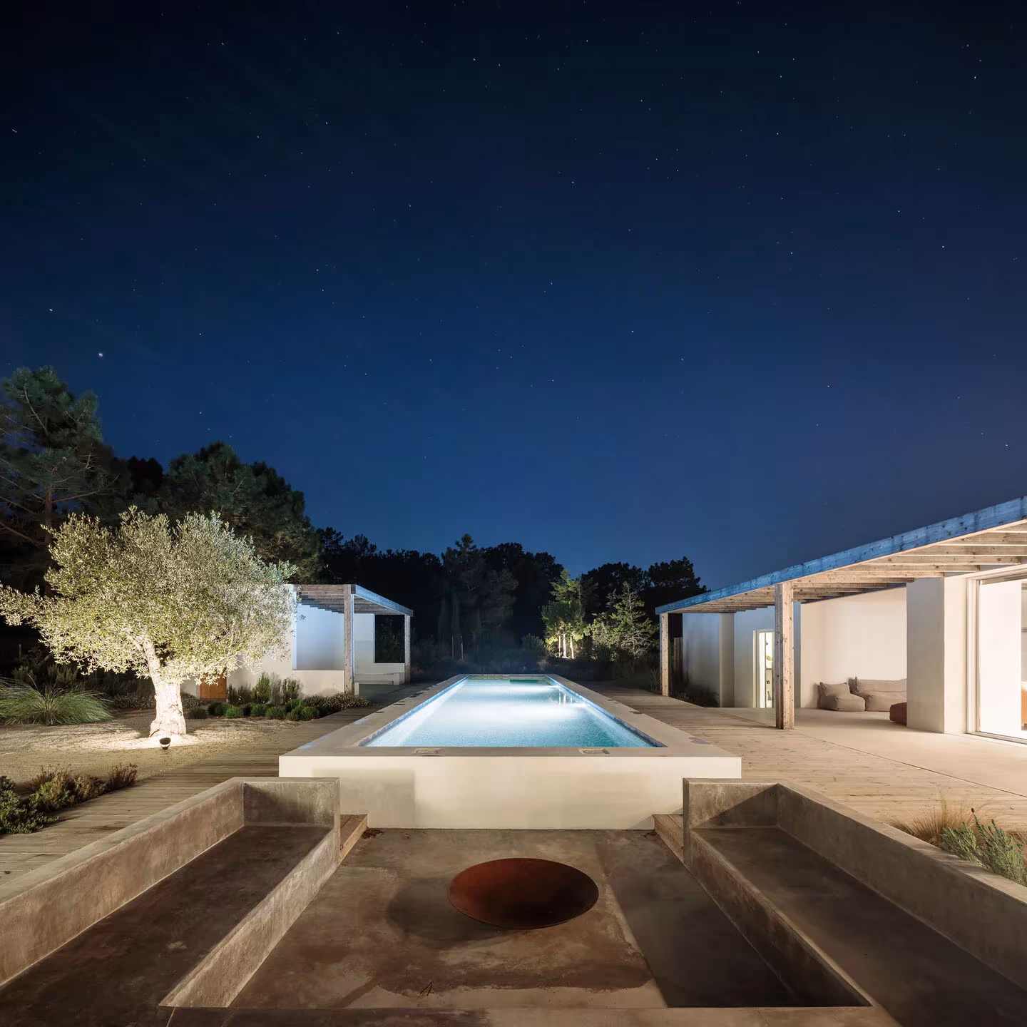 Comporta 10 de noche con la piscina iluminada, un olivo y un brasero hundido en primer plano