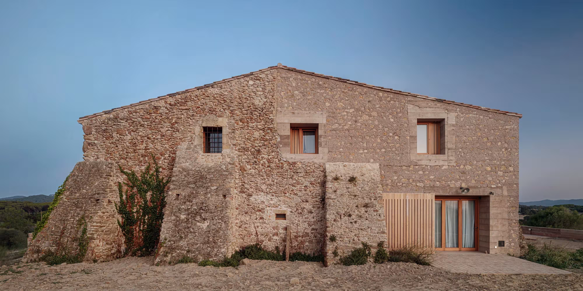 Casa 1627 de H Arquitectes, masía de piedra con carpintería de madera sobre un llano seco al atardecer