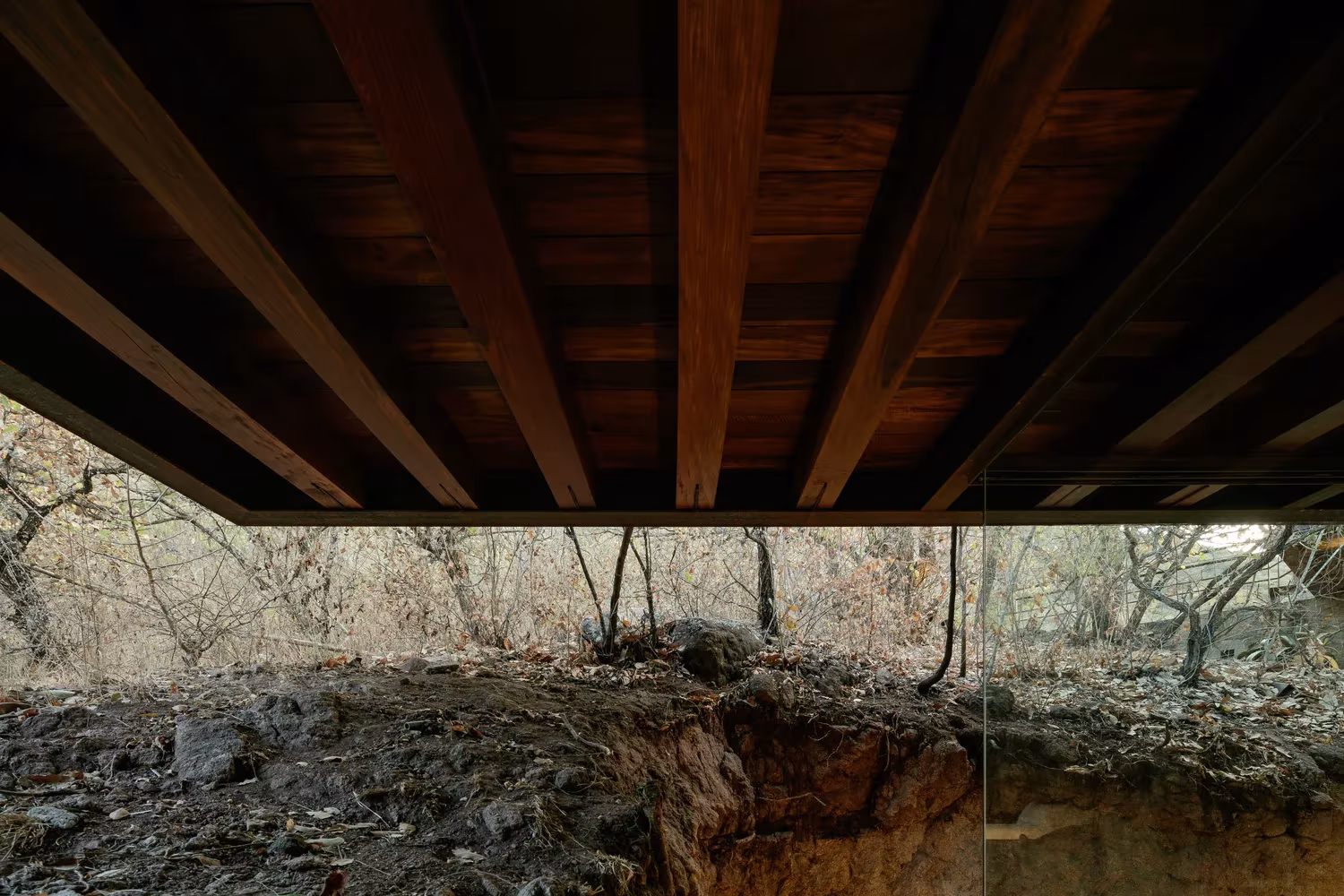 Subestructura de vigas de madera oscura del voladizo de la Cabaña Teitipac sobre el terreno de roca y raíces