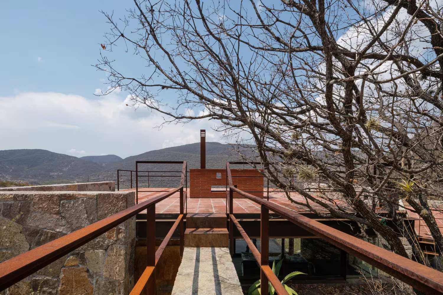 Pasarela de acero corten con barandillas que cruza hacia la cubierta de la Cabaña Teitipac y las montañas