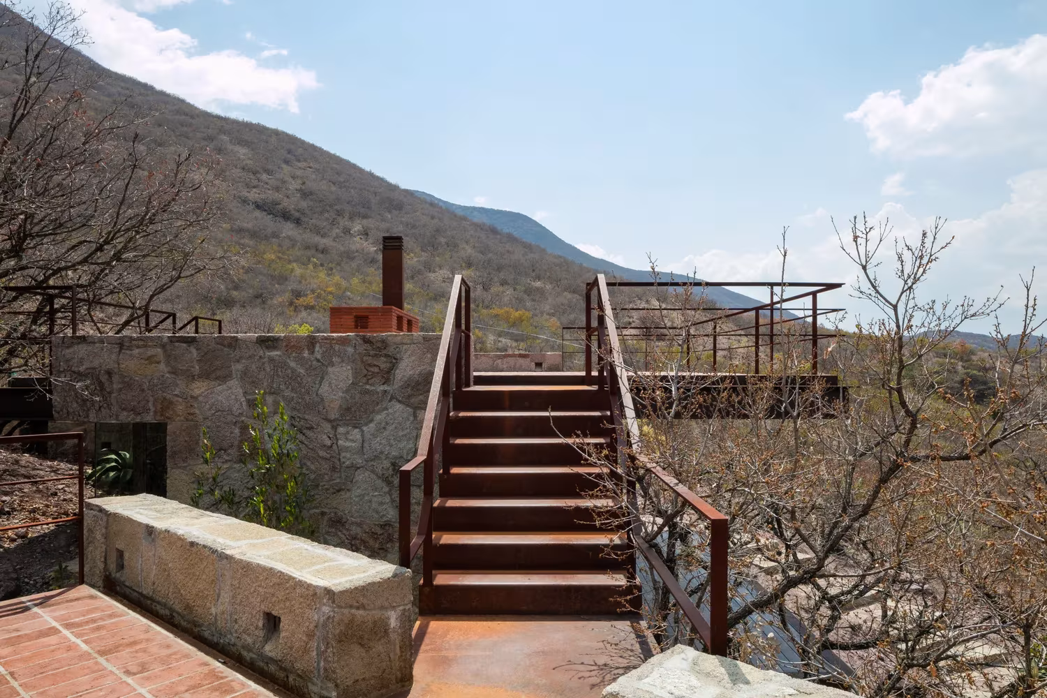 Escalera metálica de acero corten junto a un muro de piedra que sube a la azotea de la Cabaña Teitipac