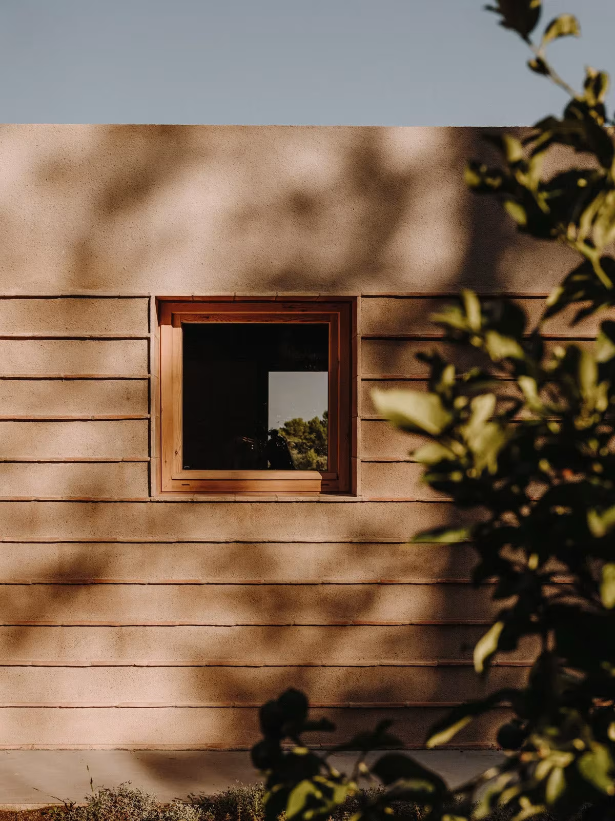 Muro de piedra de río en hiladas de la Casa Ter con ventana cuadrada de madera y sombras de hojas