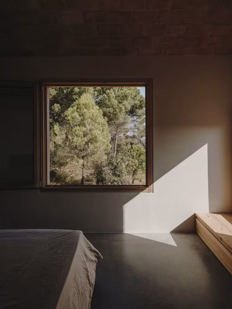 Dormitorio en penumbra de la Casa Ter con ventana cuadrada que enmarca los pinos y luz sobre la pared