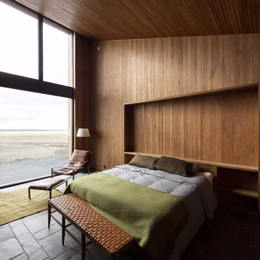 Dormitorio revestido de madera con cama de manta verde y gran ventanal a la pampa en Morro Chico