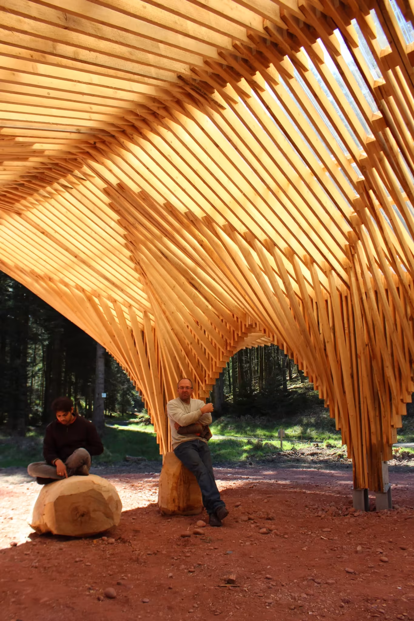 Pilares de madera arqueados y listones bajo la cubierta, con dos personas sentadas en bloques de tronco