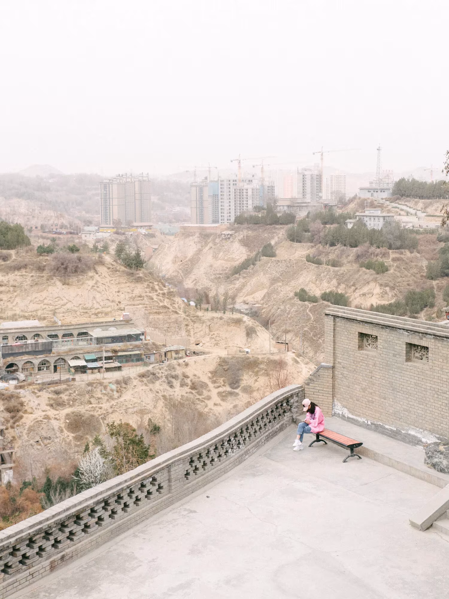 Mirador con balaustrada ornamentada sobre un barranco y una ciudad con grúas entre la bruma