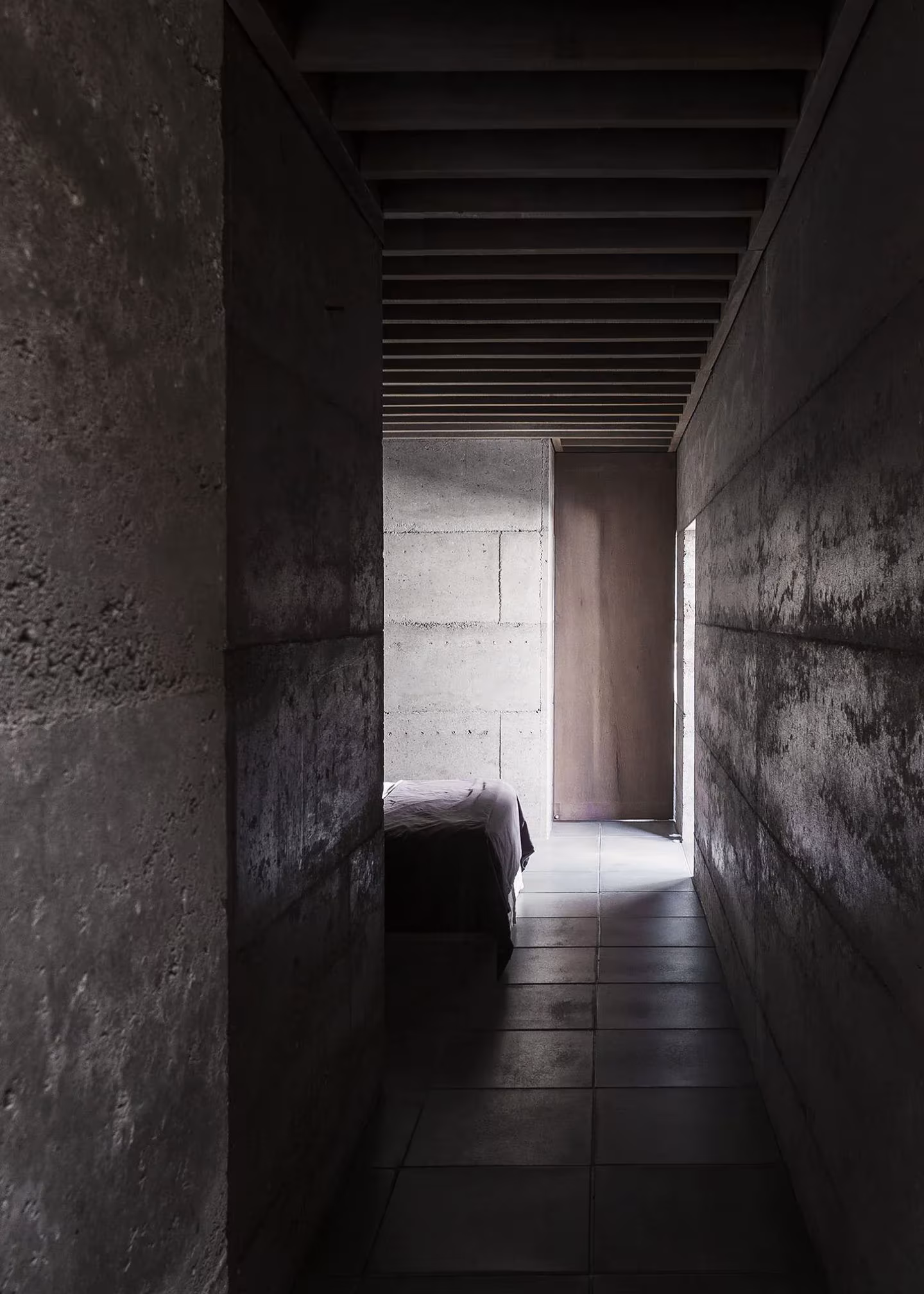 Pasillo estrecho de hormigón en penumbra que lleva a un dormitorio en Cloister House
