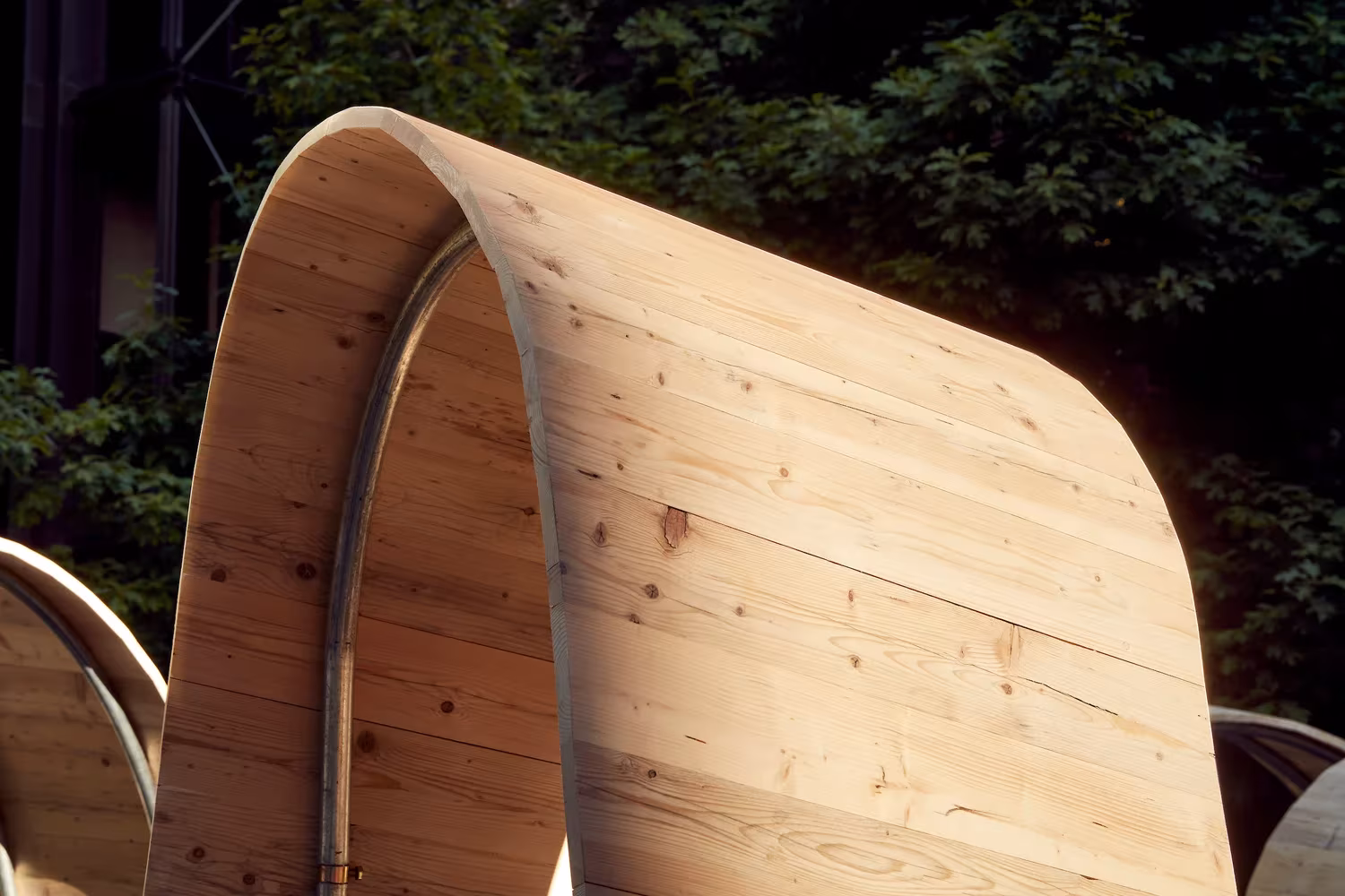 Detalle de una curva de madera laminada de Please Be Seated, de Paul Cocksedge, entre árboles