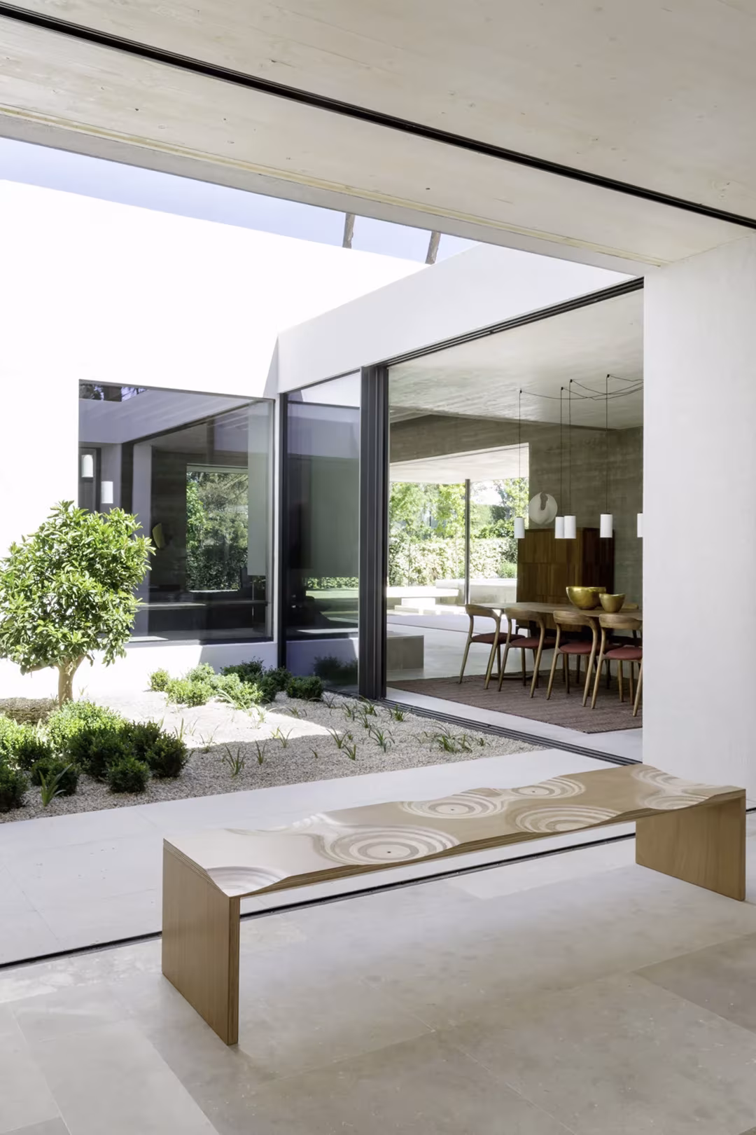 Patio interior con árbol y banco de madera ante el comedor acristalado del Proyecto AS de ÁBATON