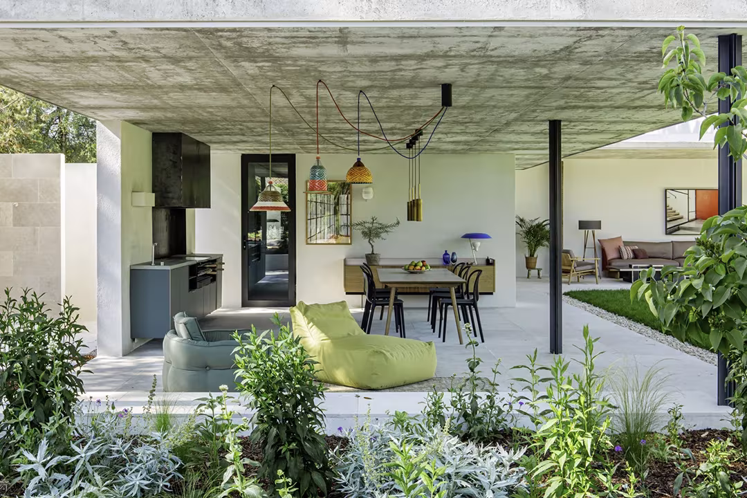 Porche de hormigón visto con cocina exterior y comedor abiertos al jardín, Proyecto AS de ÁBATON