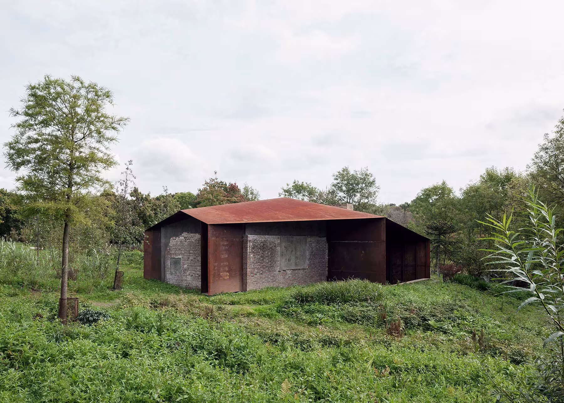 Pabellón de acero corten de Carmody Groarke entre prados y árboles, con cubierta inclinada sobre muros de ladrillo
