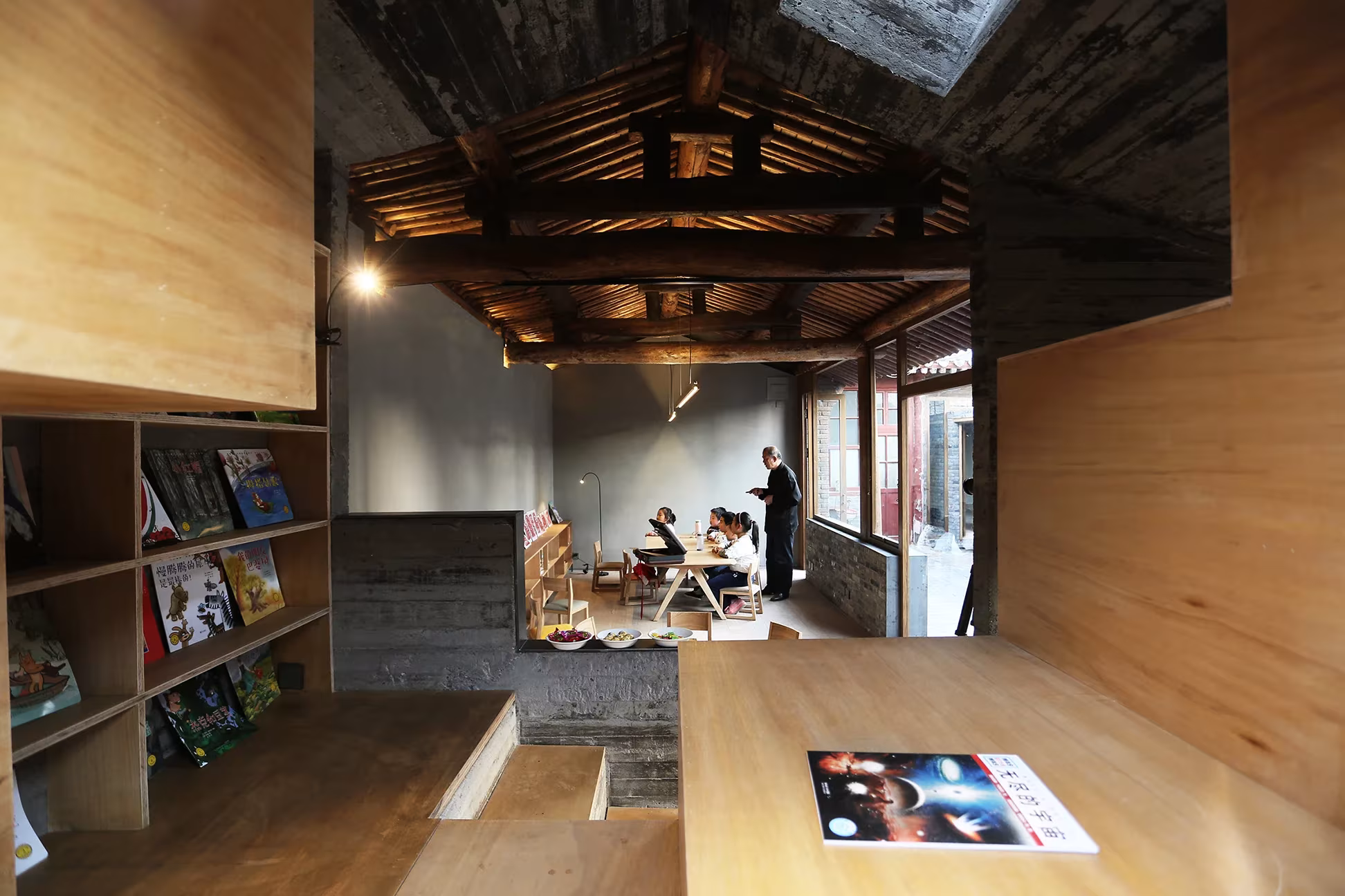 Sala de lectura con vigas de madera vistas, estanterías de libros infantiles y mesa larga en el hutong