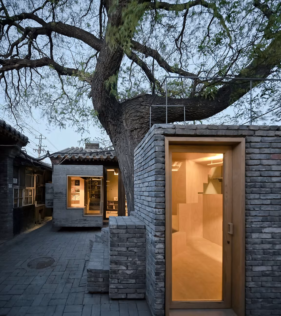 Puerta de madera clara en un volumen de ladrillo gris bajo un gran árbol del hutong de Pekín