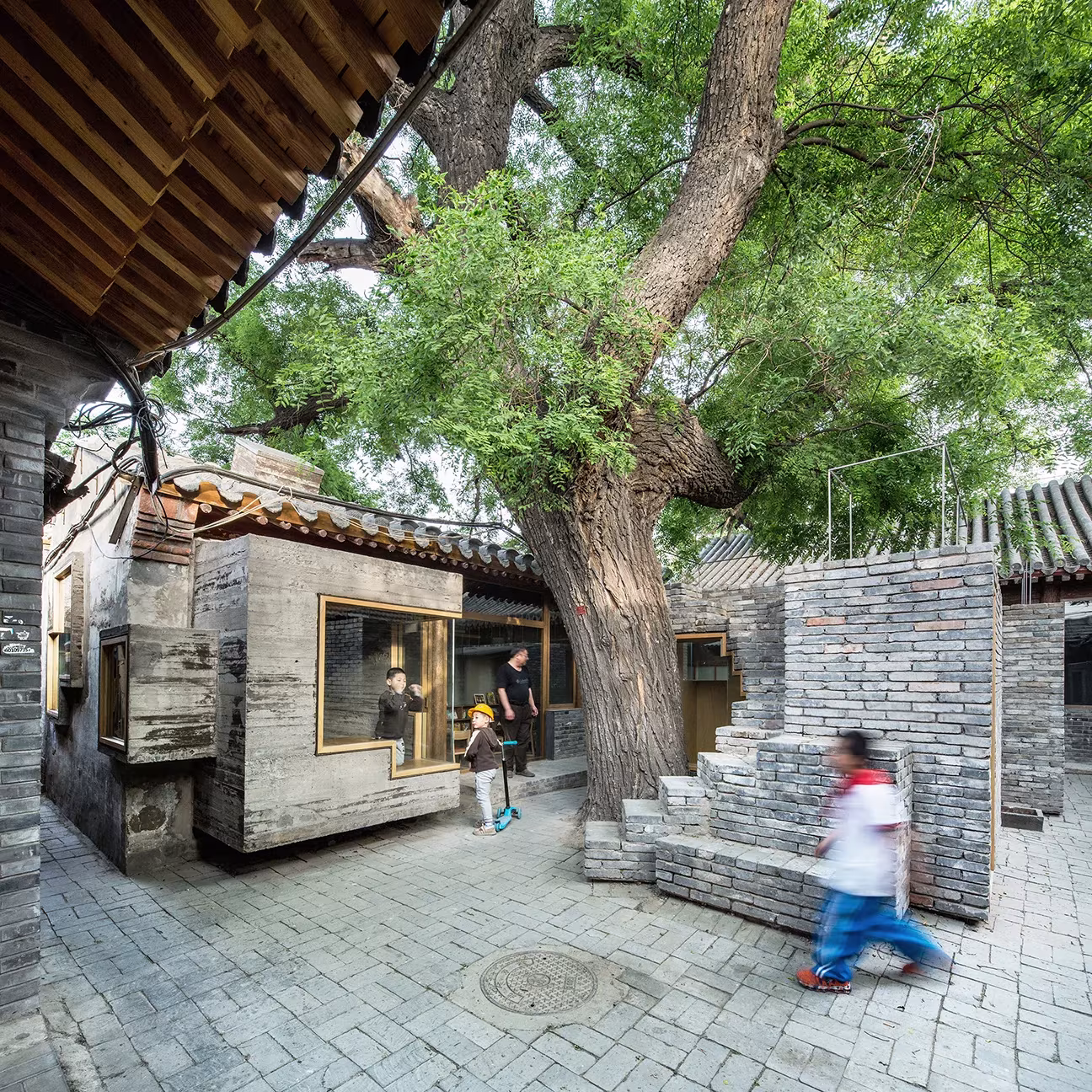 Niños corriendo en el patio con escalera de ladrillo y árbol grande de la minibiblioteca del hutong