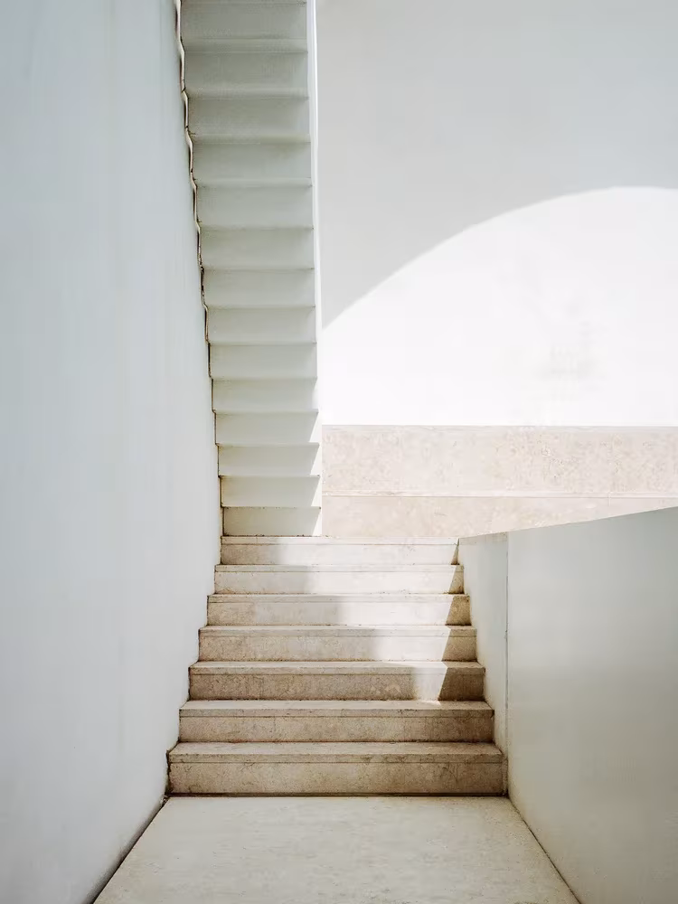 Escalera de piedra clara entre muros blancos con luz de arriba en el estudio de Aires Mateus, Lisboa