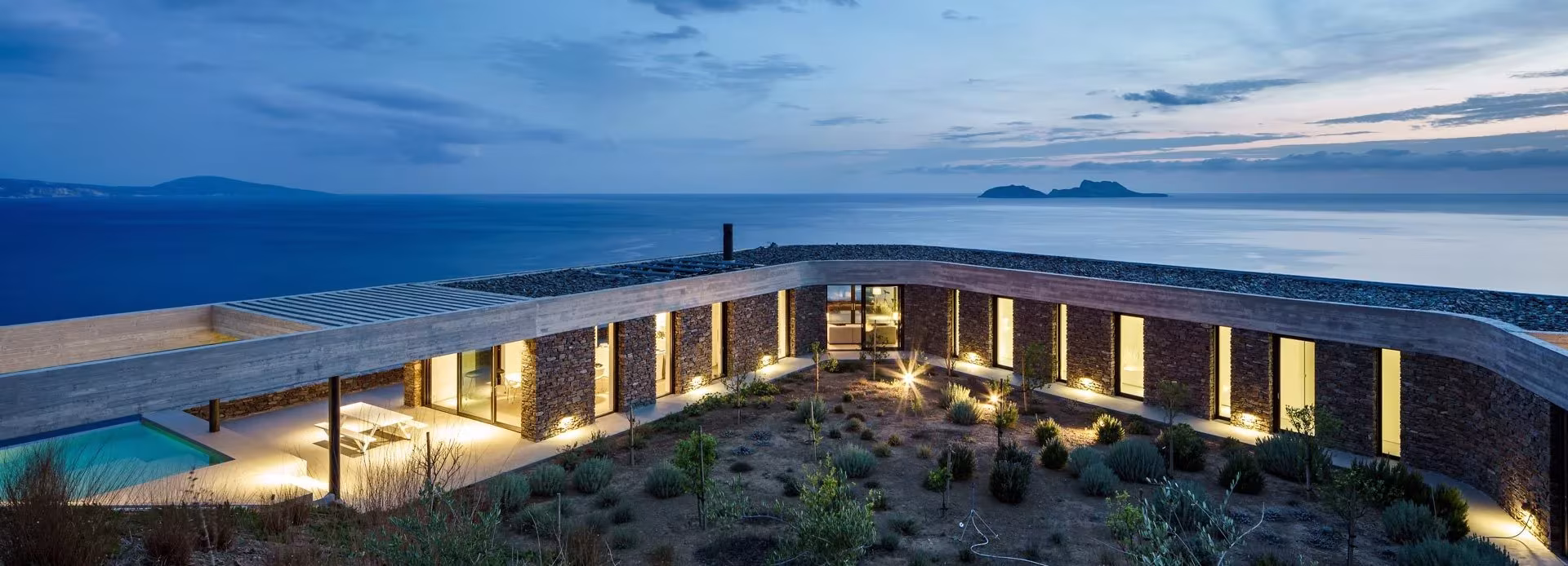 Ring House de DECA Architecture al anochecer, con el patio interior iluminado, la piscina y el mar al fondo