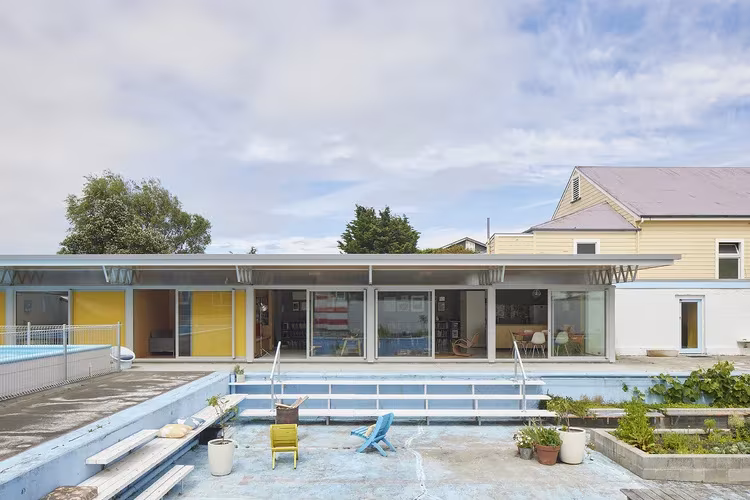 Gonville Pool House: la antigua piscina pública con un edificio bajo de cubierta voladiza y carpinterías amarillas