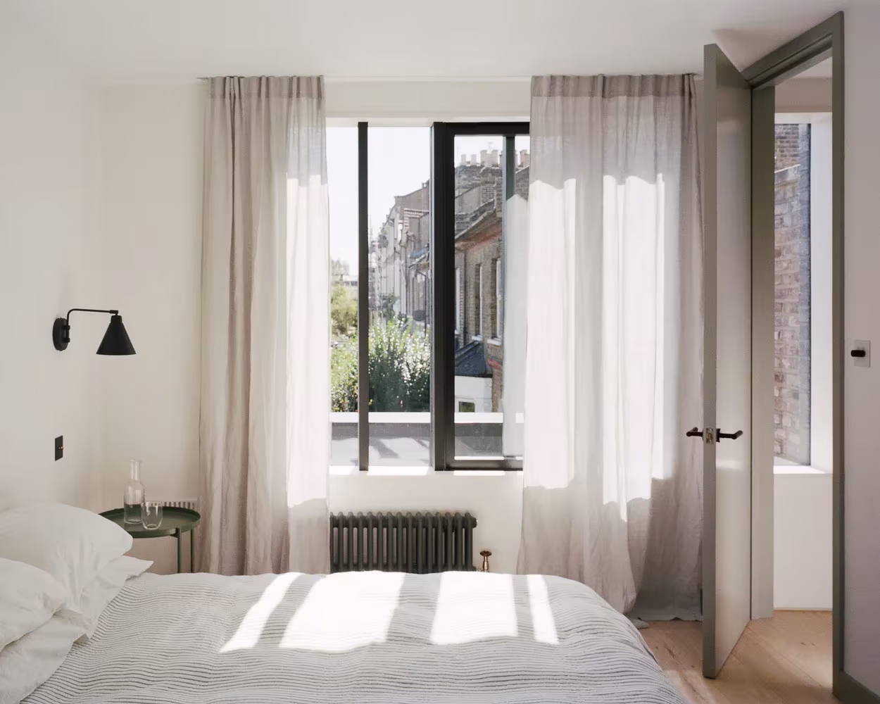 Dormitorio blanco de Corner House con cortinas de lino y ventana de carpintería negra sobre la calle
