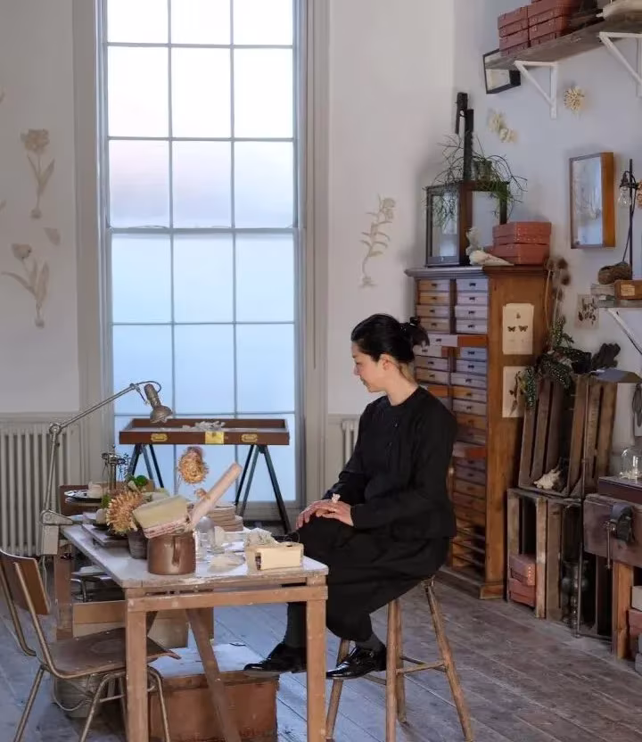Kaori Tatebayashi trabajando en su mesa de taller junto a un ventanal alto y un archivador de cajones