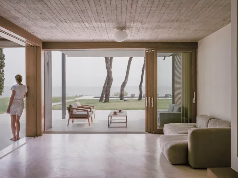 Salón de St. Minas House, de Neiheiser Argyros, con techo de madera y cristaleras abiertas al jardín y al mar