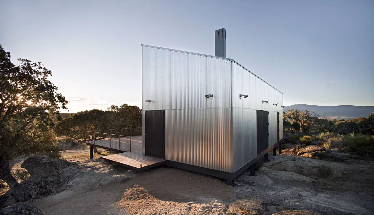 Casa Garoza elevada sobre pilotes metálicos entre rocas y encinas de la sierra de Ávila, al atardecer