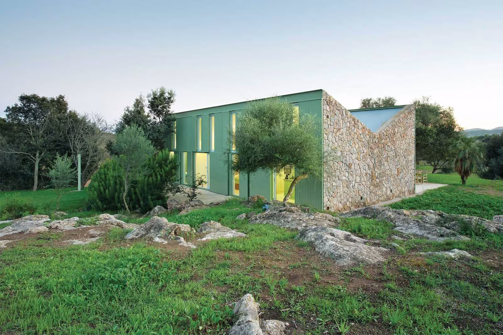 Fachada verde con ventanas amarillas de la Casa Cobo junto a muros de piedra en un prado con rocas