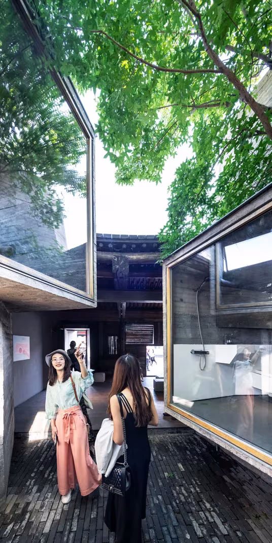 Dos visitantes en el patio de Micro Hutong, entre cajas acristaladas que asoman bajo las copas de los árboles
