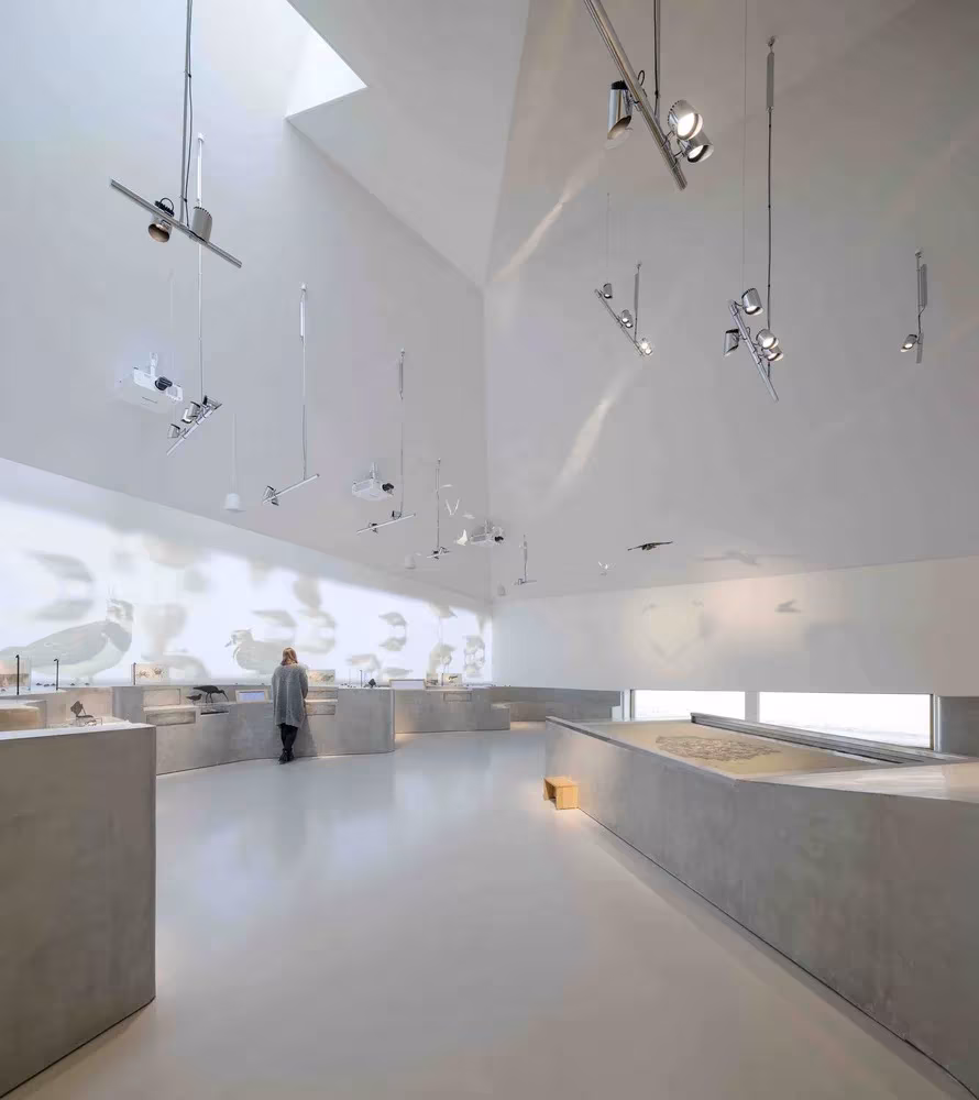 Sala expositiva blanca de techo facetado con mostradores de hormigón en el Centro del Mar de Wadden de Dorte Mandrup