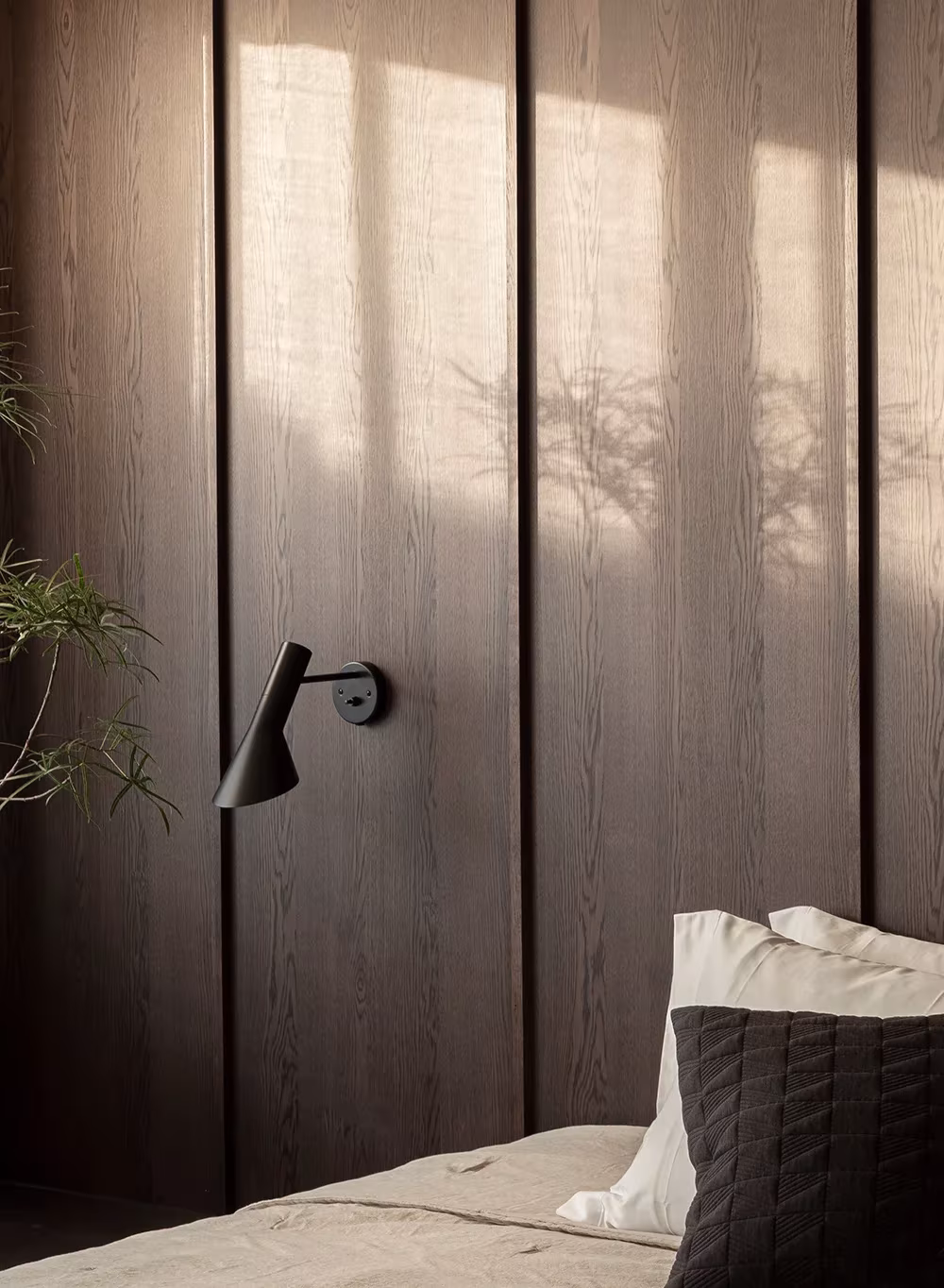 Panelado de madera oscura con luz rasante, aplique negro y sombras de plantas junto a la cama en Azabu, Tokio