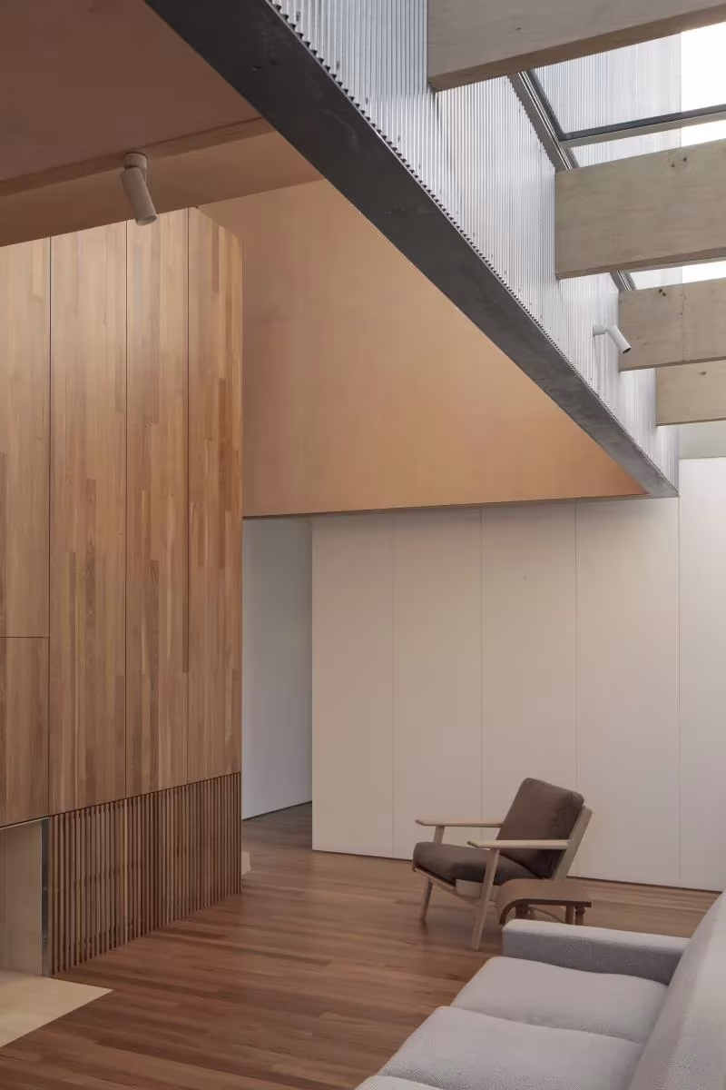 Interior de la Casa AB con revestimiento de madera, lucernario, butaca y sofá bajo una pasarela