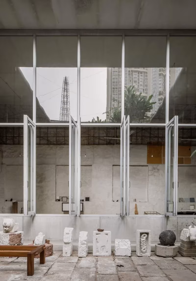 Muro de carpinterías metálicas abatibles sobre un patio con maquetas y piezas de piedra en Studio Mumbai, Alibag