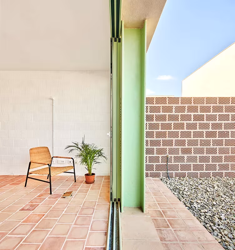 Terraza con suelo de baldosa de barro, butaca, planta y carpintería verde en las viviendas VPO de Inca