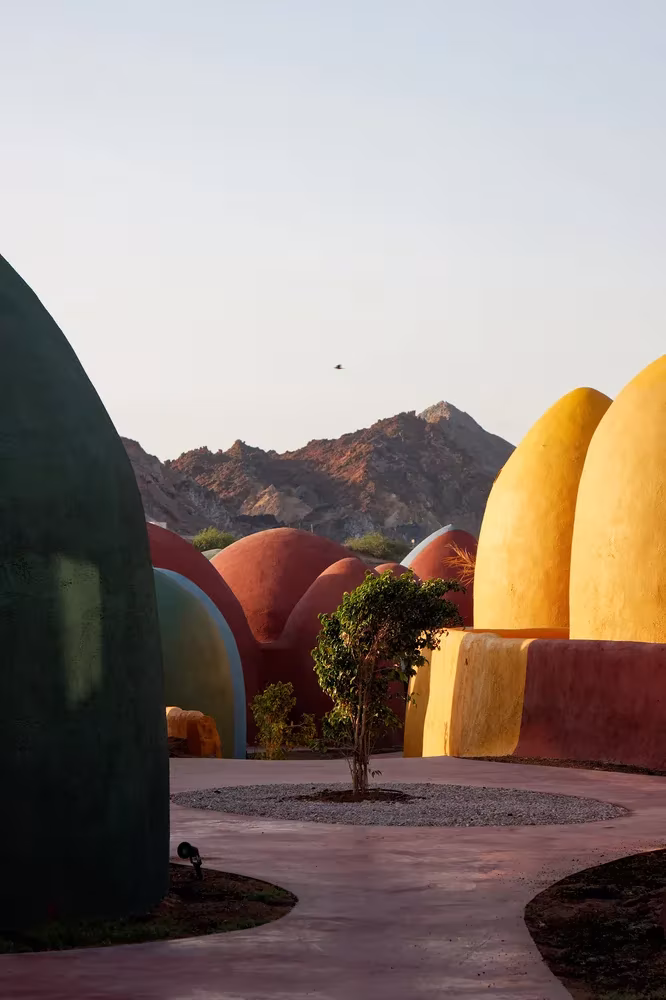 Cúpulas de adobe amarillas y rojas ante la montaña, con un árbol en el patio del poblado de Ormuz