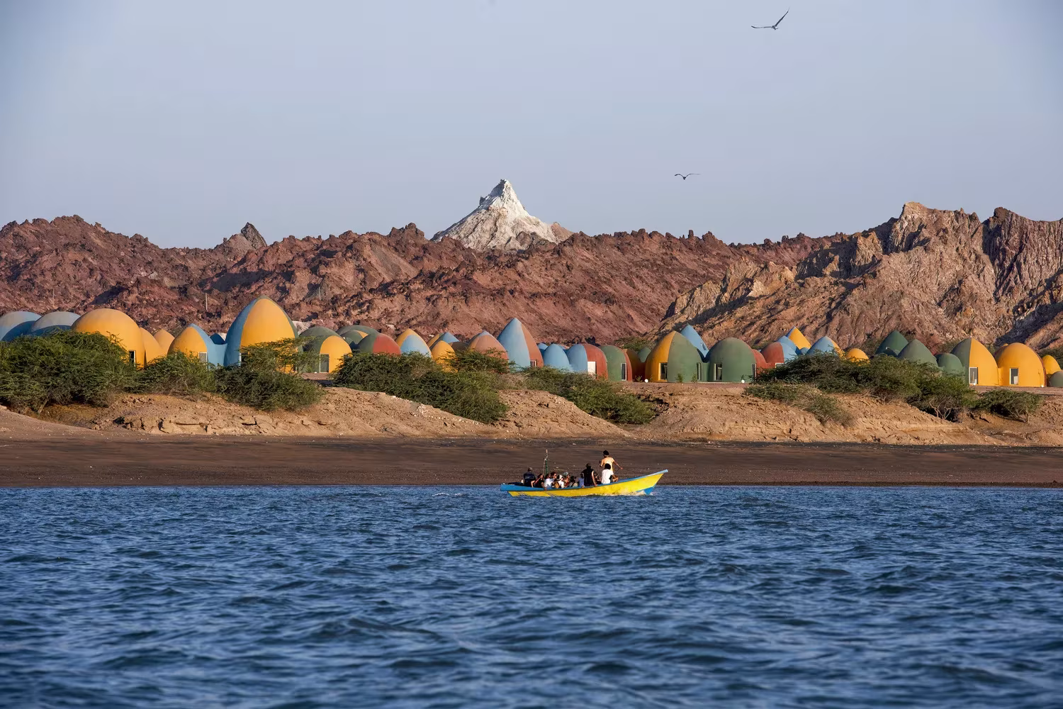 Poblado de cúpulas de colores junto al agua, con una barca y montañas áridas al fondo en Ormuz