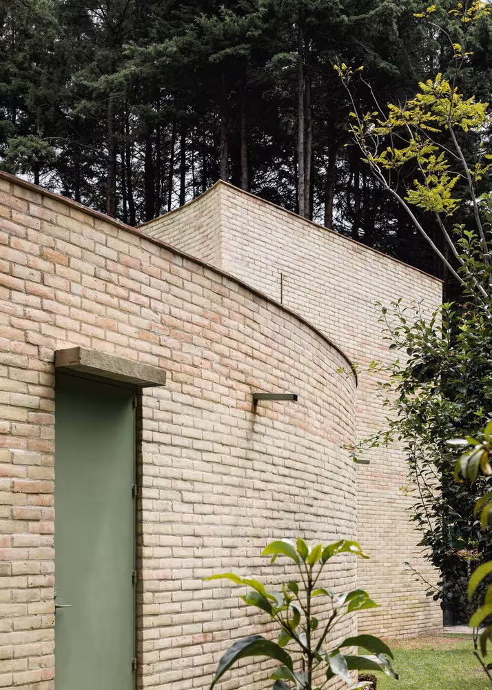 Muro de ladrillo curvo con puerta verde frente al jardín en Forest House de LANZA Atelier