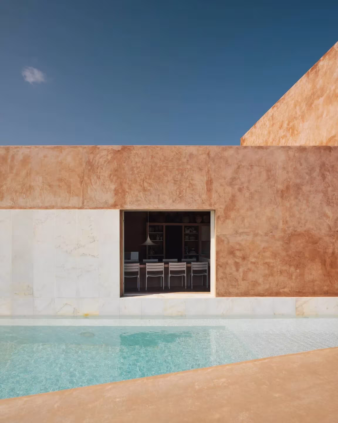 Muro de tono ocre con ventana cuadrada sobre una piscina de agua turquesa en una casa en Grândola