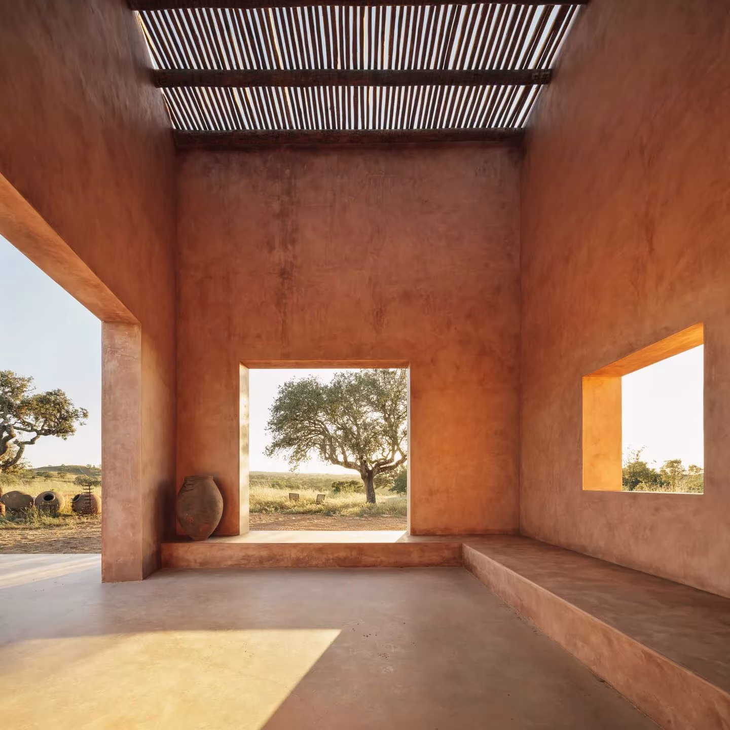 Estancia de muros terracota con pérgola y huecos cuadrados que enmarcan alcornoques en una casa en Grândola