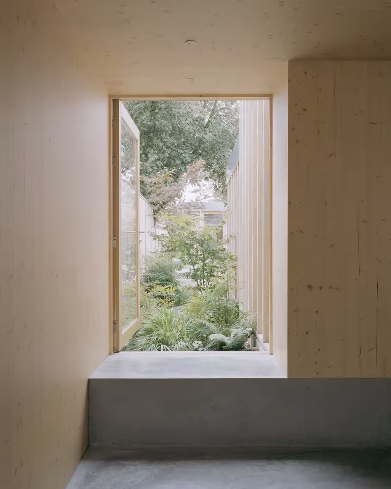 Rincón de madera clara en la Casa y estudio Spruce con banco de hormigón bajo una ventana al jardín