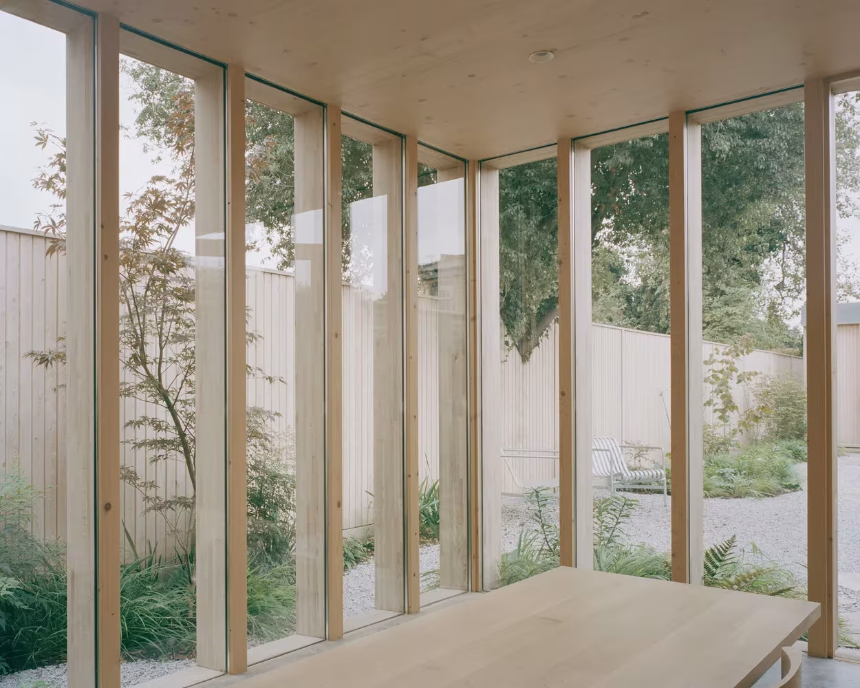 Estancia acristalada de la Casa y estudio Spruce con mesa de madera y vistas a un patio con árboles