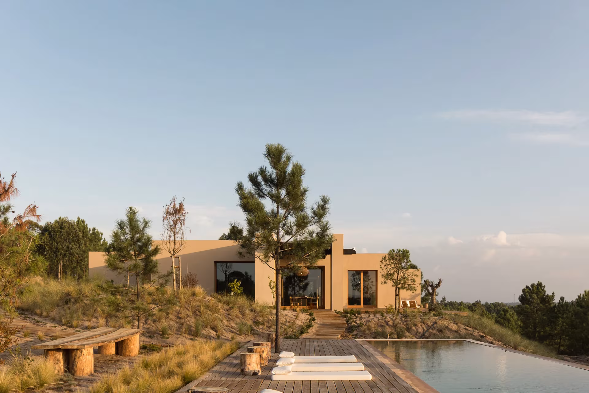 Melides Art entre pinos y matorral, con bancos de madera junto a una piscina desbordante al atardecer