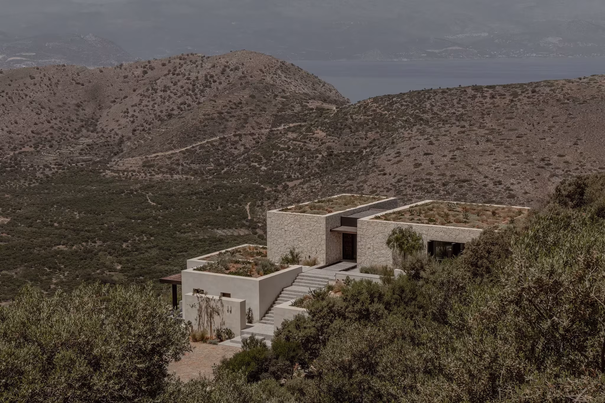 Casa O Lofos de piedra y cubiertas planas en una ladera de matorral mediterráneo frente a montañas
