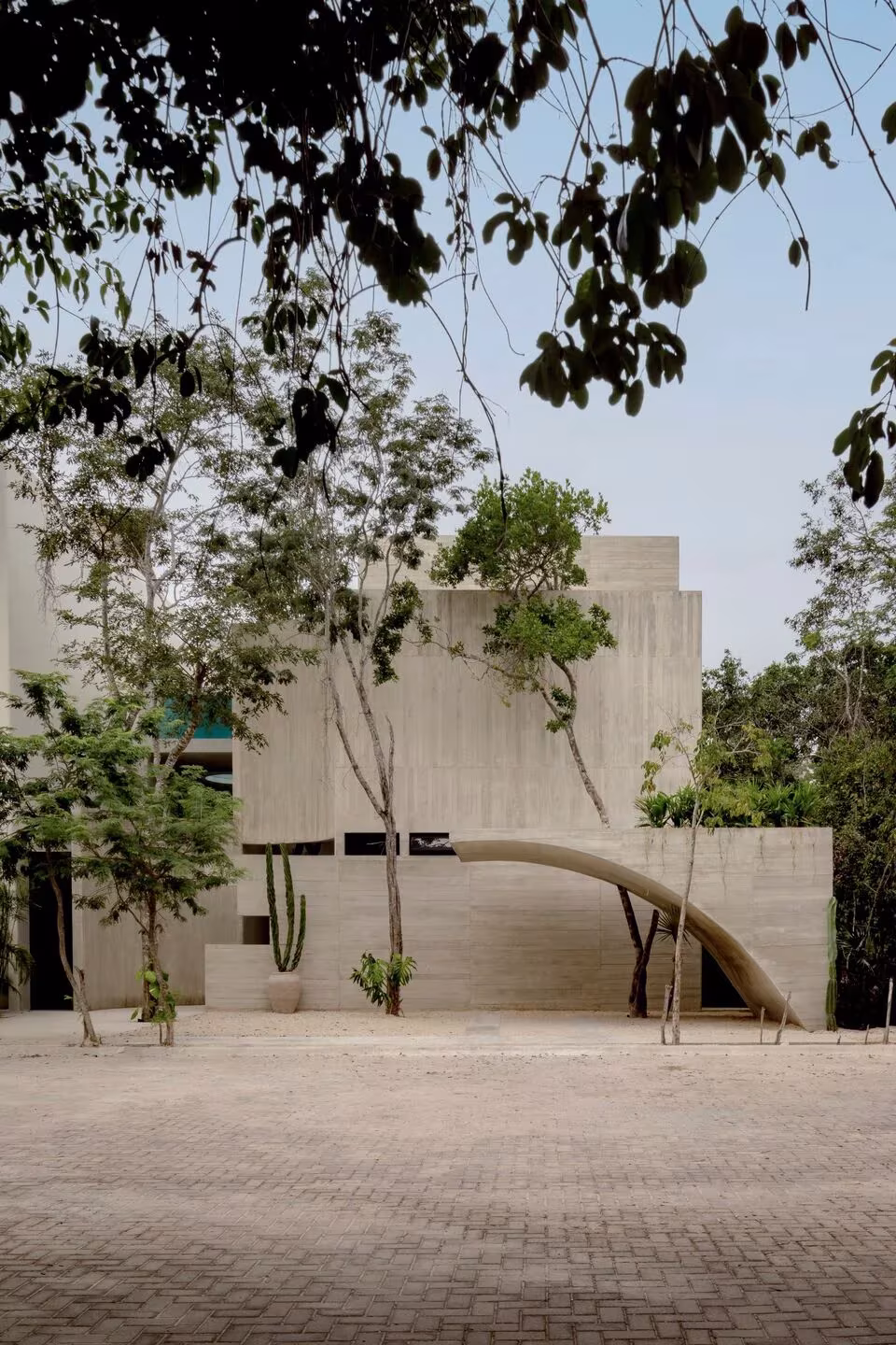 Villa Cava de hormigón claro entre árboles, con un arco rebajado sobre un patio adoquinado