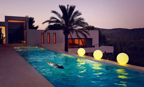 Long swimming pool at dusk with glowing spheres, a palm tree and a lit house beyond