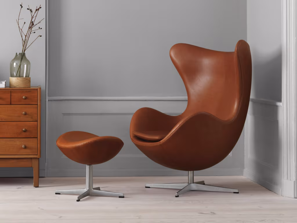 Arne Jacobsen Egg chair in brown leather with a footstool, beside a timber chest of drawers on a grey wall