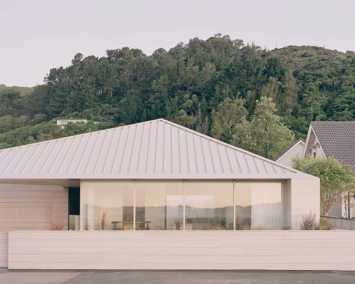 RK Residence, casa baja con cubierta metálica gris y gran ventanal, frente a una ladera arbolada
