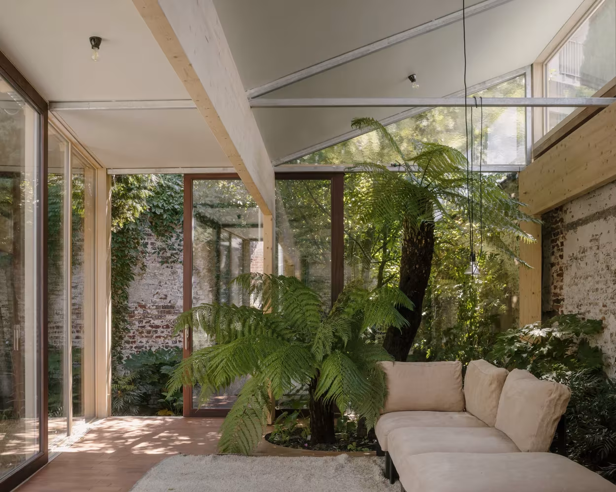 Salón de Martelaar House con sofá claro entre helechos y muros de ladrillo, bajo vigas de madera